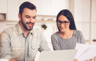 Smiling young man using a laptop sitting next to a young woman holding papers. Both are looking at the laptop screen.